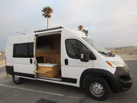 White RV rental camper van with an open side door, showing a custom wood interior, parked near a sandy beach with palm trees.
