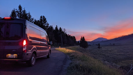 A dark RV rental van shines with a vibrant sunset reflection, parked on a forest road overlooking a vast valley.