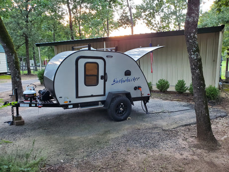 A compact Bushwhacker teardrop camper, ideal for an RV rental, is parked at a wooded campsite with its hatch open.