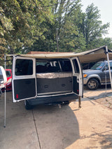 This white camper van, an ideal RV rental, has its rear doors open revealing a bed setup. An attached awning offers shade.