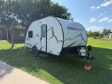 A white Freesolo travel trailer, an rv rental, parked on a sunny green lawn next to a house. Features a distinct mountain graphic.
