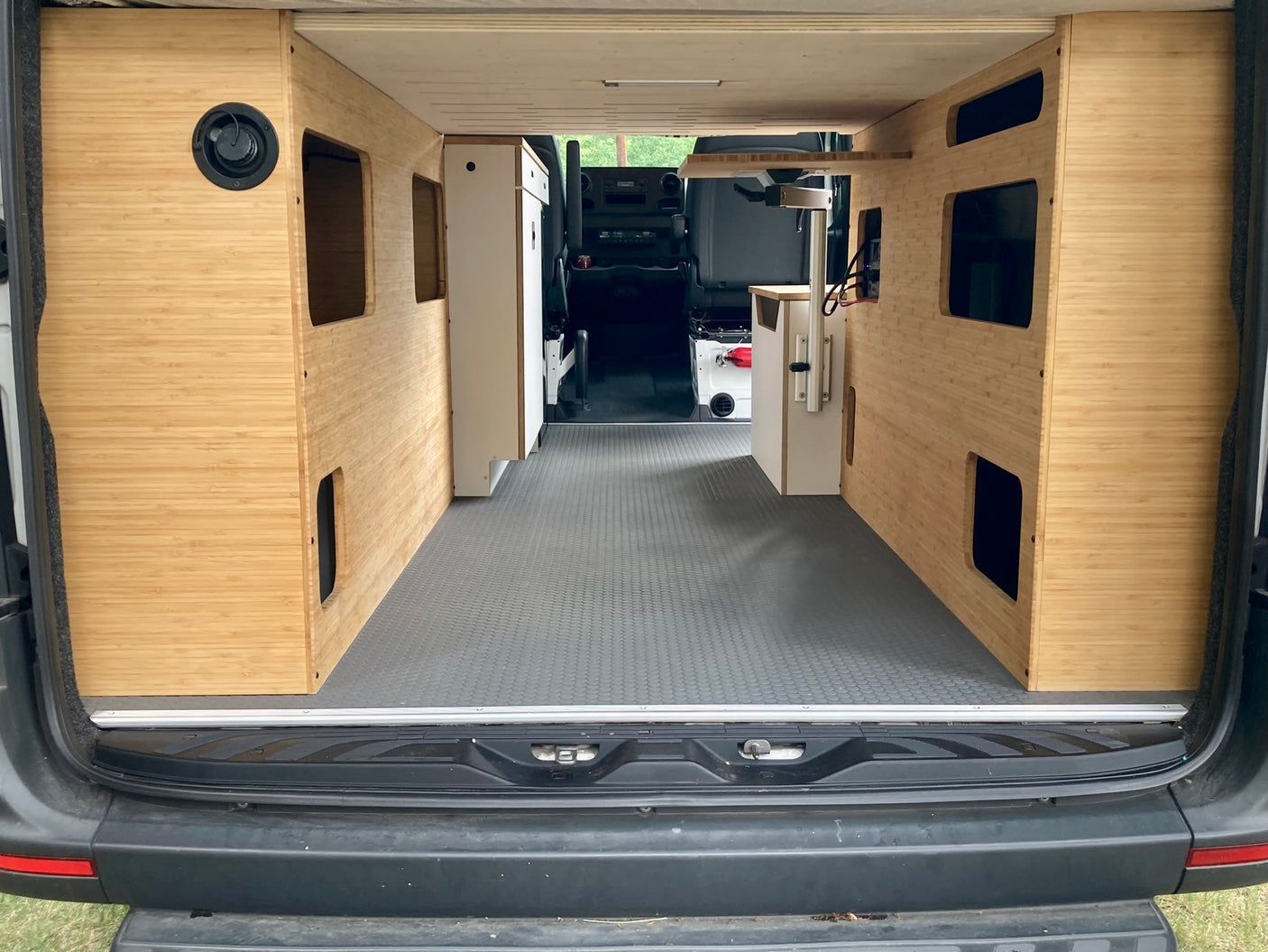 Modern RV rental van interior with custom light wood panels, built-in storage, and grey floor, viewed from the rear.