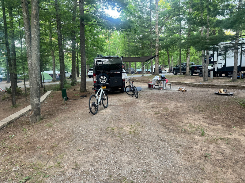 Campsite with a black van-style rv rental, bikes, picnic table, grill, and fire pit under trees. — photo 13