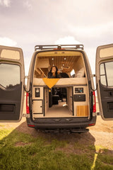 Relax in your spacious RV rental! A woman enjoys the custom camper van's cozy interior with rear doors open to nature.
