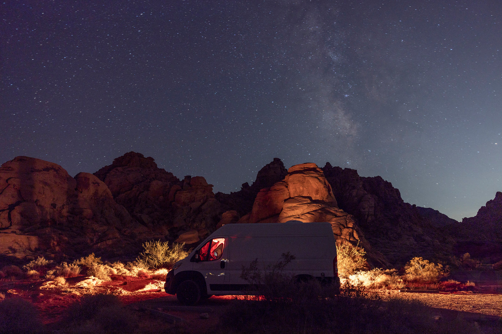 White RV rental van with red interior light, parked in a desert landscape under the starry Milky Way.