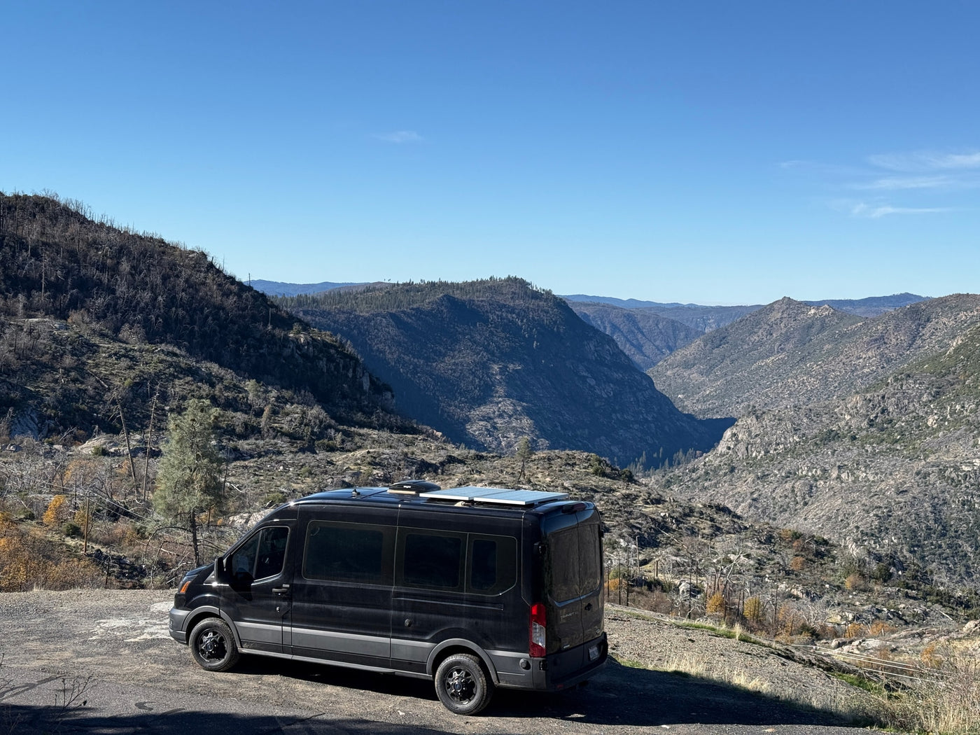 A black camper van (RV rental) parked on a dirt road overlooking a vast, rugged mountain valley with a clear blue sky. — photo 13