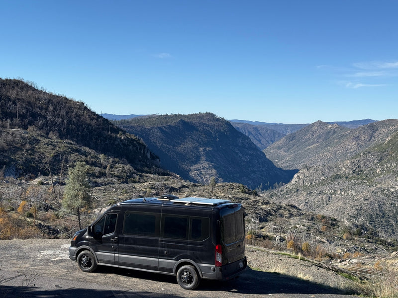 A black camper van (RV rental) parked on a dirt road overlooking a vast, rugged mountain valley with a clear blue sky. — photo 13