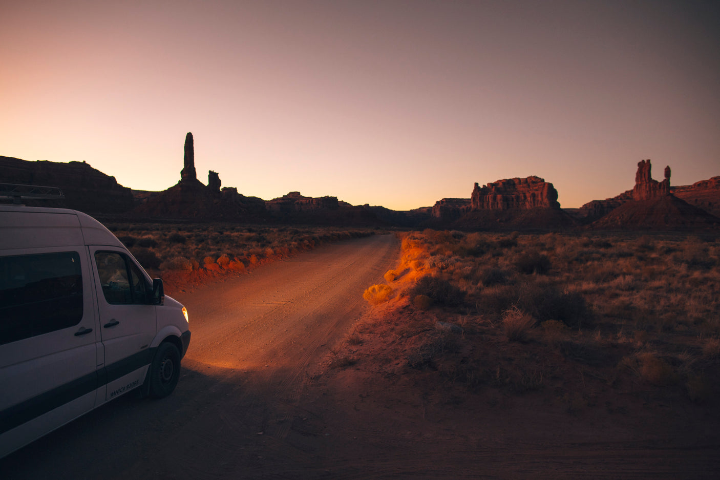 An RV rental van on a dirt road, its headlight on, illuminating the vast desert. Towering rock formations loom at sunset.