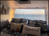 Cozy RV rental interior with bed & pillows. Stunning ocean view from the back, showing a distant sailboat.