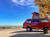 Explore by rv rental: Red camper van parked on a sandy beach by the ocean under a clear blue sky, pop-top deployed.