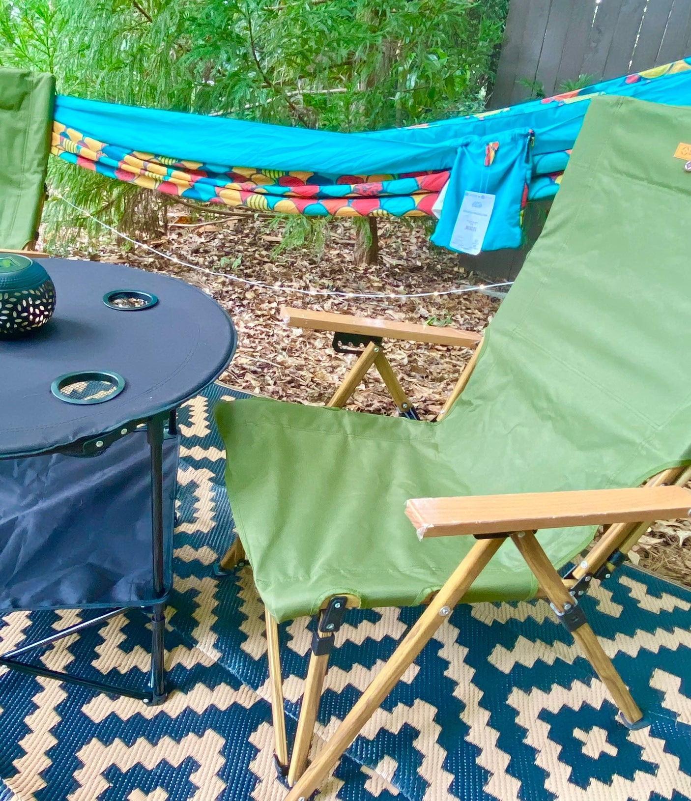 Cozy outdoor leisure area for your RV rental getaway. Features a colorful hammock, green camping chairs, and table on a patterned rug.