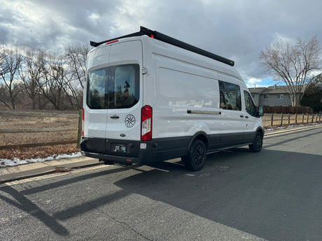 White Ford Transit camper van for RV rental, parked outdoors. Features roof rack, awning & compass decal.