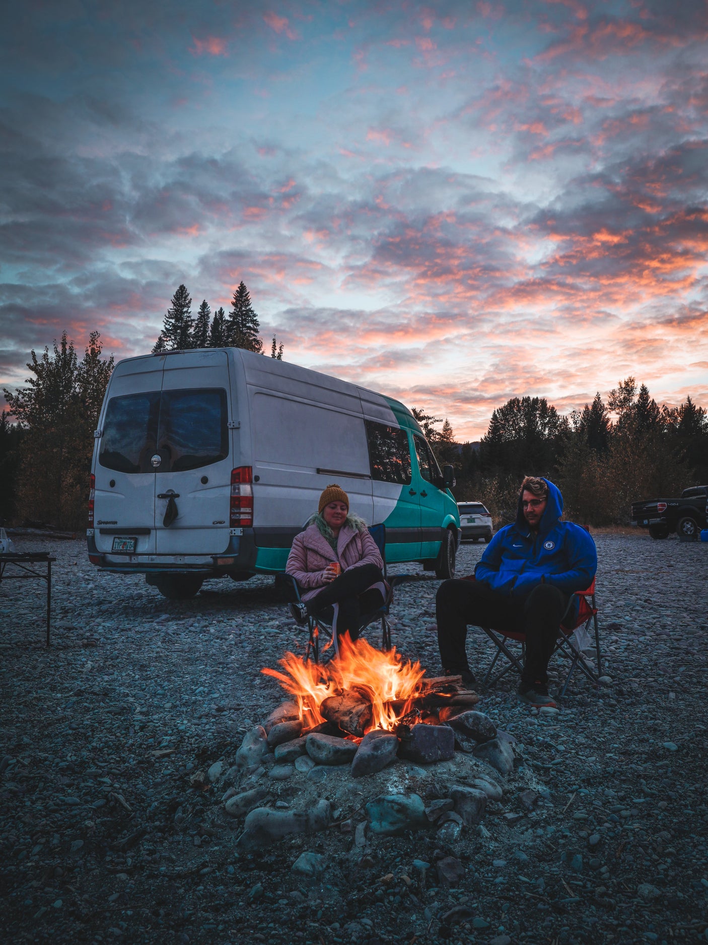Two people relaxing by a warm campfire at sunset, beside their **RV rental** van on a gravel riverbed. Ideal for nature trips.