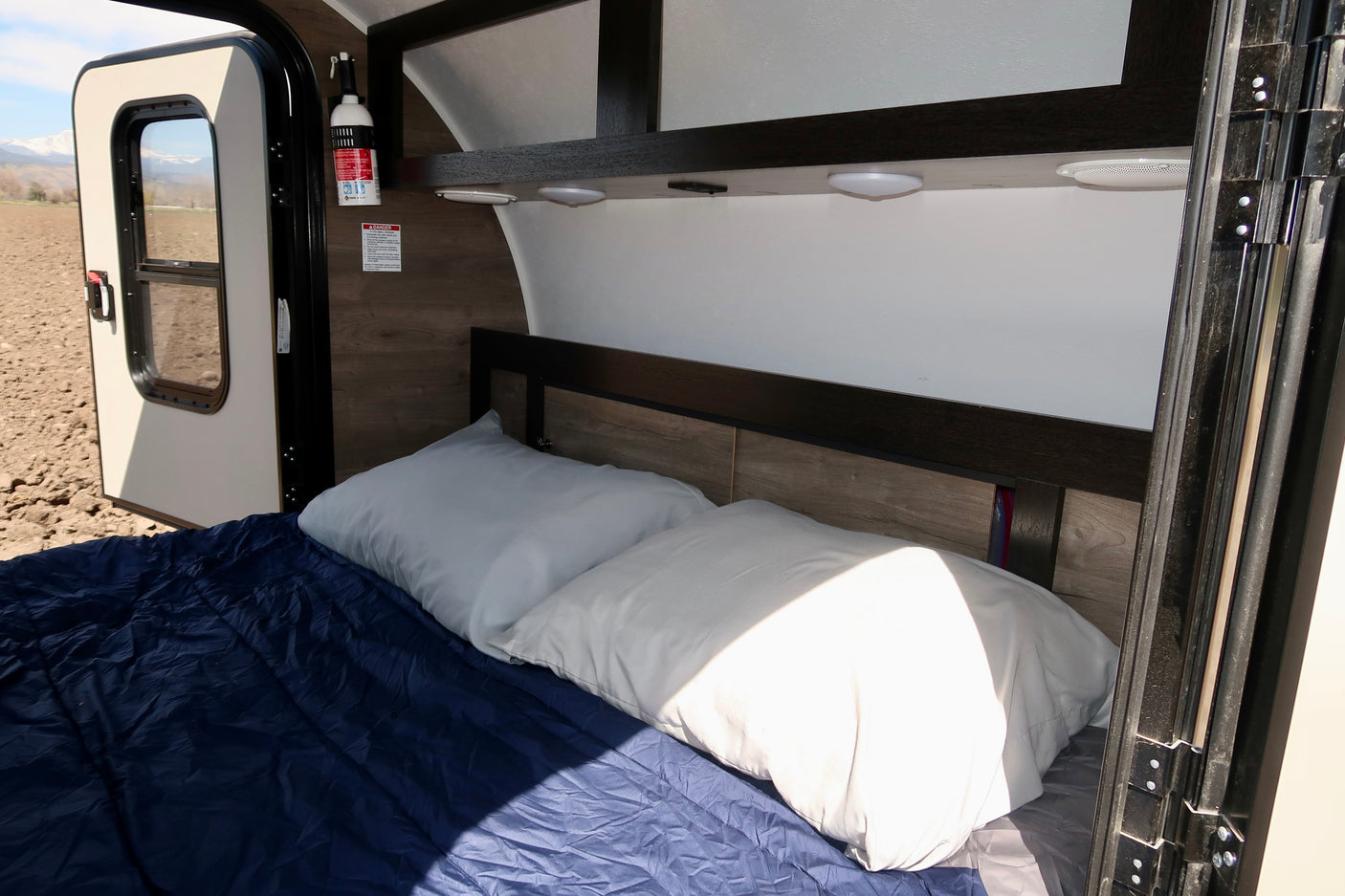 Cozy RV rental interior: bed, pillows, and a blue blanket. Open door reveals mountain views. Fire safety on wall.