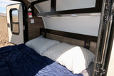 Cozy RV rental interior: bed, pillows, and a blue blanket. Open door reveals mountain views. Fire safety on wall.