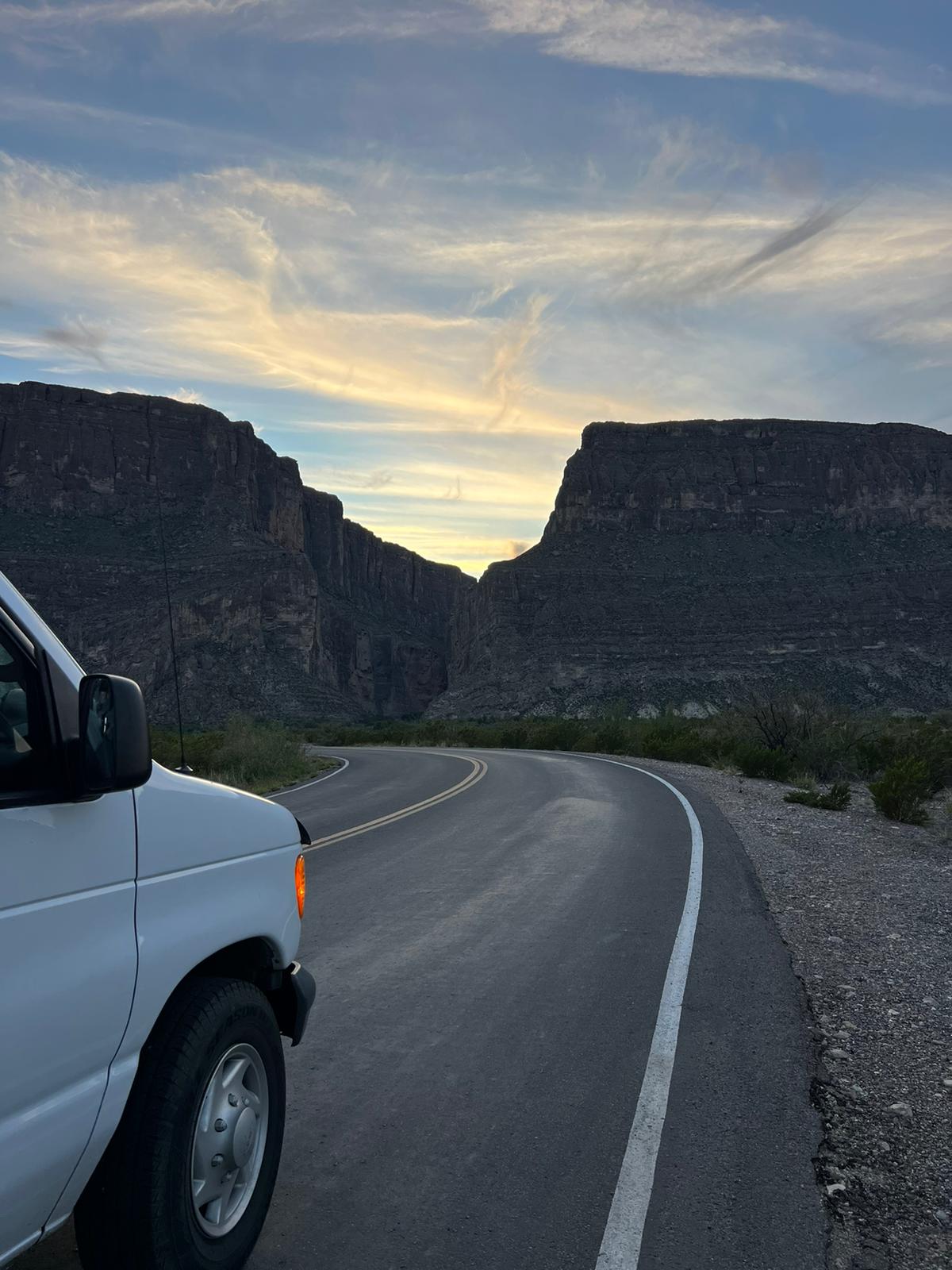 A white RV rental on a curving road, leading into a grand canyon between towering cliffs under a dramatic sunset sky. Perfect road trip.