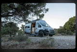 Silver camper van (RV rental) parked in a dry, natural landscape with trees. Its side door is open, revealing the wooden interior.