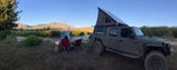 Scenic RV rental: a customized Jeep Gladiator with a pop-top camper parked by a campfire and river in a mountainous landscape.
