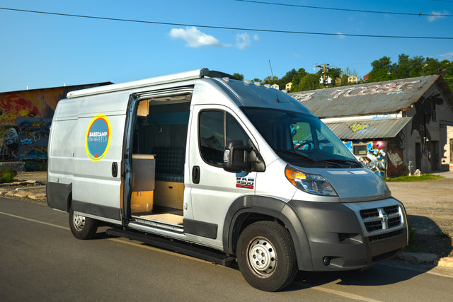 Silver Ram ProMaster camper van, an RV rental with "Basecamp on Wheels" logo, parked with its side door open revealing the custom interior.