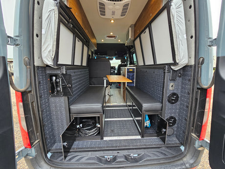 Step into this spacious RV rental camper van interior, featuring rear-facing bench seating, a dining table, overhead cabinets, and practical storage.