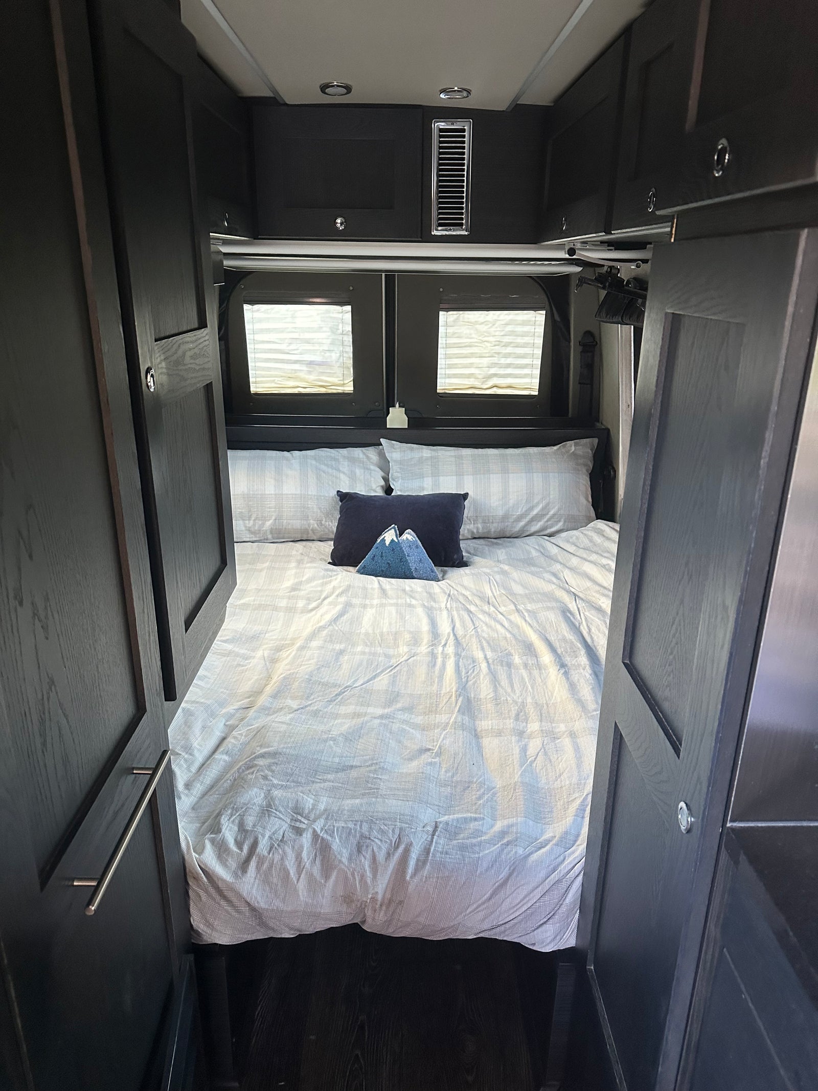 Step into this comfortable RV rental bedroom. Features a large bed with striped bedding, dark cabinets, and rear windows.
