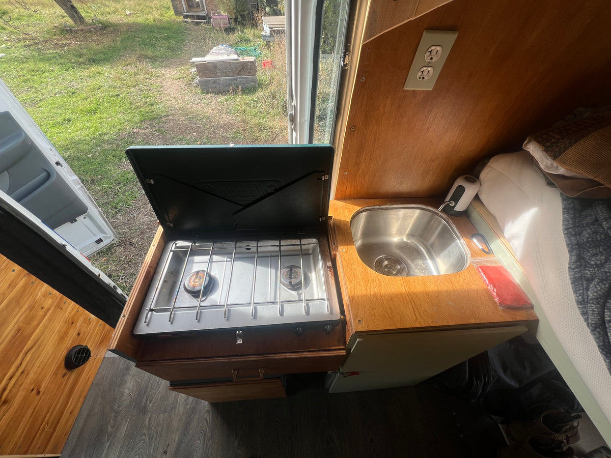 Compact rv rental kitchen features a 2-burner stove, sink, and nearby bed. Enjoy the outdoor view from this functional camper space. — photo 5