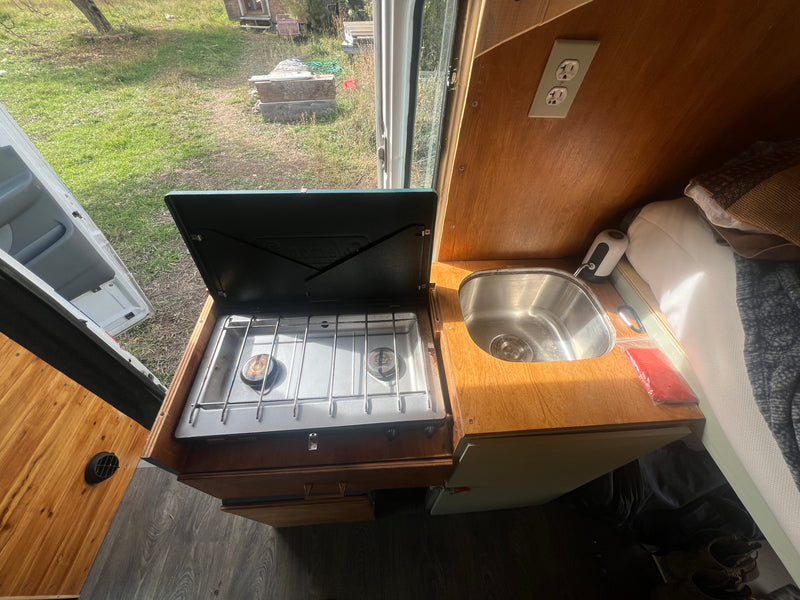 Compact rv rental kitchen features a 2-burner stove, sink, and nearby bed. Enjoy the outdoor view from this functional camper space. — photo 5
