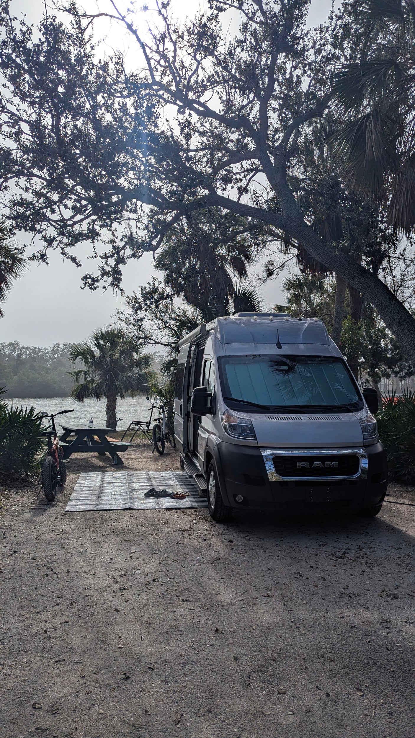 Grey rv rental van parked at a scenic waterfront campsite with bikes, picnic table, and palm trees for outdoor adventure.
