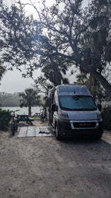 Grey rv rental van parked at a scenic waterfront campsite with bikes, picnic table, and palm trees for outdoor adventure.