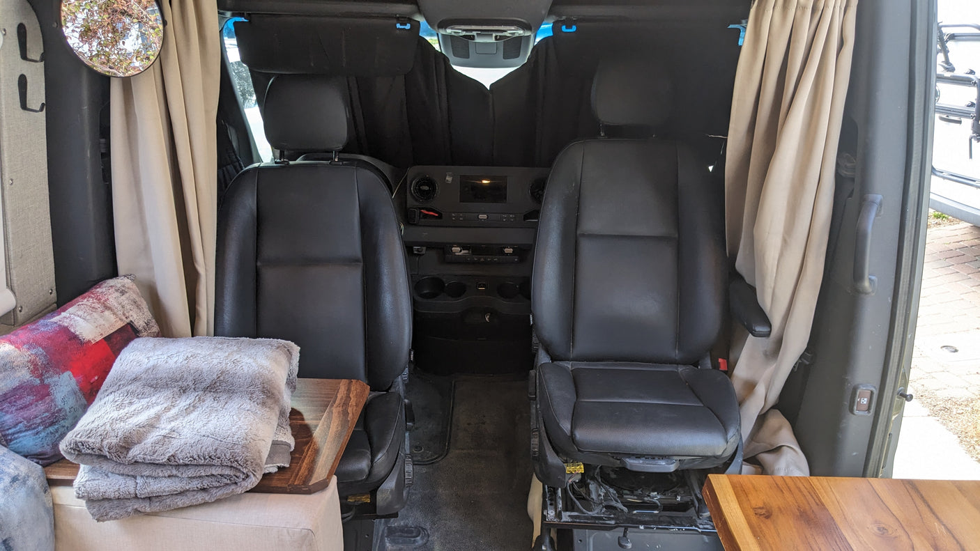 Interior of an RV rental: swiveling front seats, dashboard, side curtains, and a glimpse of the cozy living area with blankets.