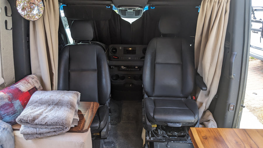 Interior of an RV rental: swiveling front seats, dashboard, side curtains, and a glimpse of the cozy living area with blankets.