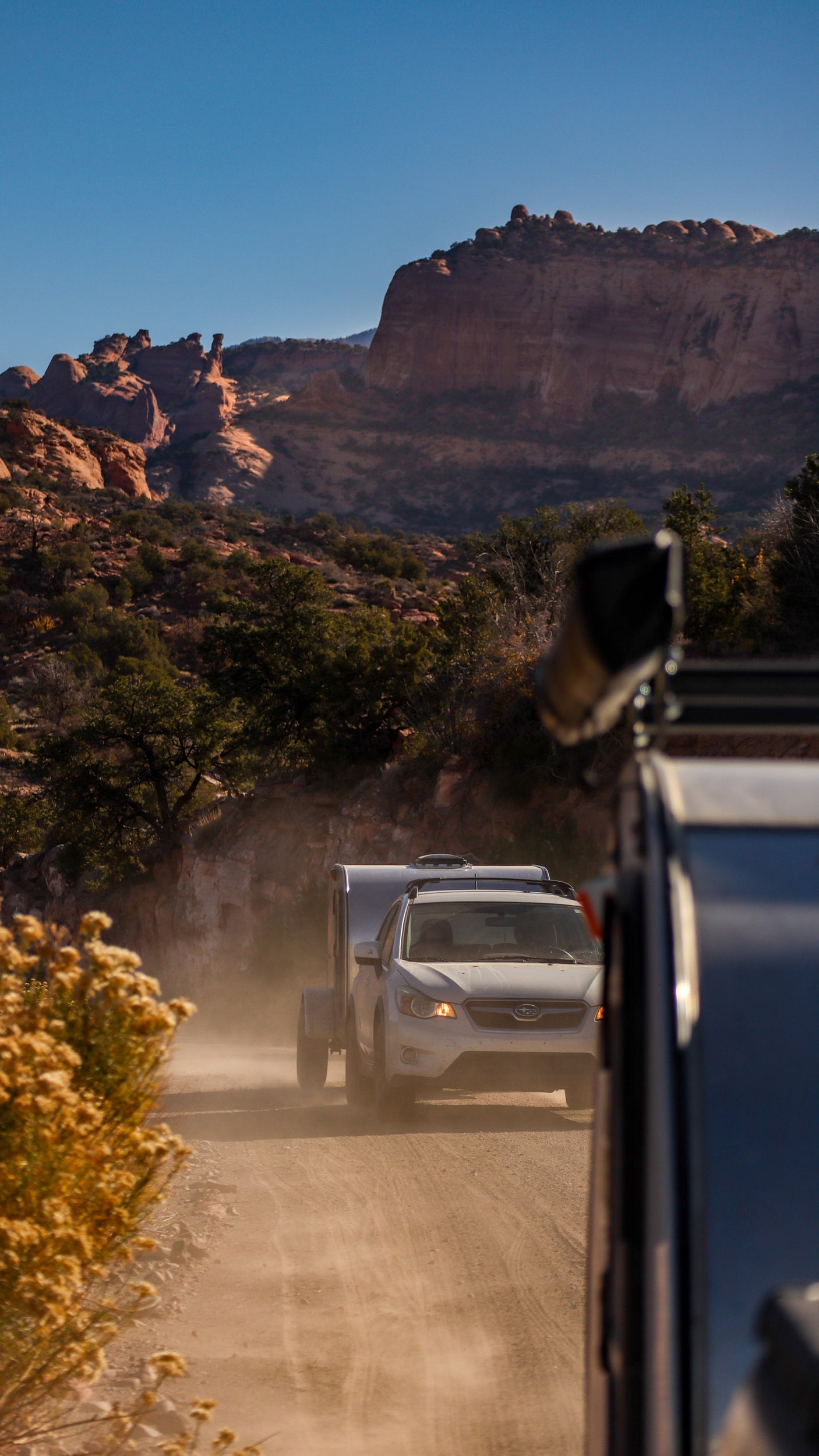 White SUV with an RV rental trailer kicks up dust on a dusty desert road through red rock mountains.