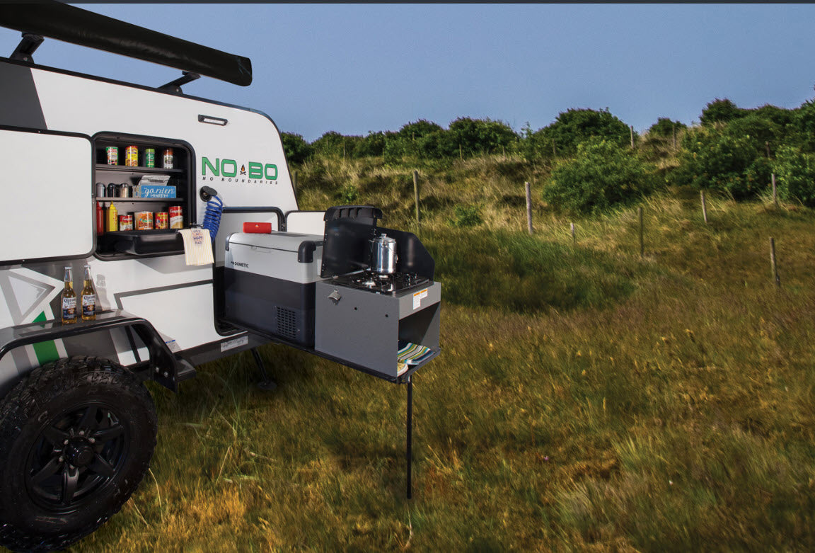 Travel trailer RV rental features an outdoor kitchen with stovetop & fridge, perfect for remote camping in a scenic grassy field.