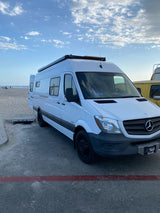 White Mercedes Sprinter rv rental camper van parked by a sandy beach under a blue sky, ready for adventure.
