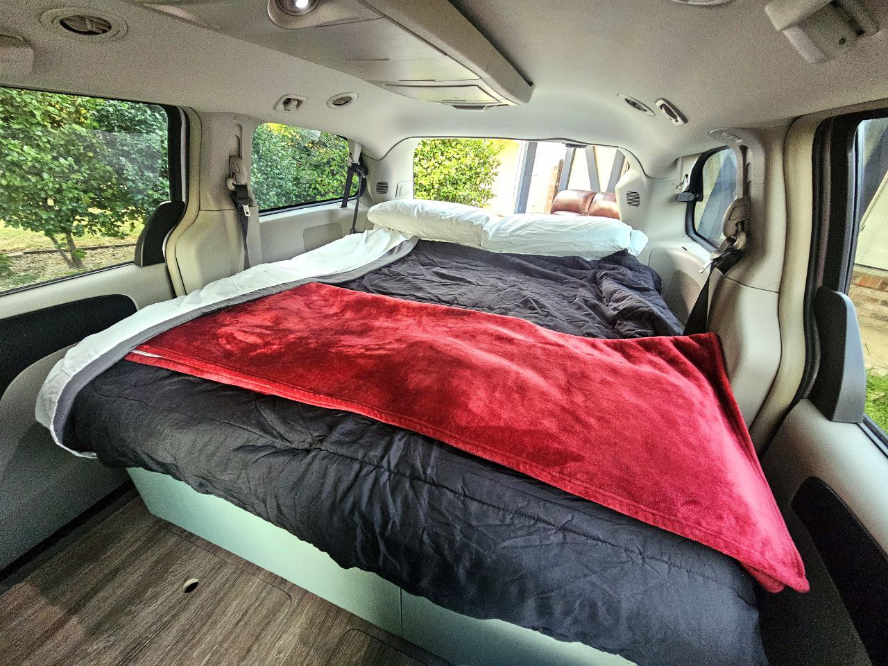 Inside an RV rental conversion: a cozy minivan bedroom with a black comforter, red blanket, and wood floor. — photo 14