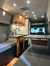 Inside a stylish RV rental van conversion: a cozy kitchen with wood cabinets, hexagon tiles, and comfy gray seating. Bright interior!
