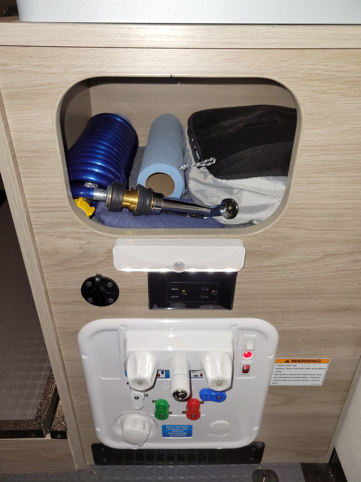 Essential RV rental utility panel: water valves, power, solar port, and storage with hose & shower sprayer. Includes safety warnings.