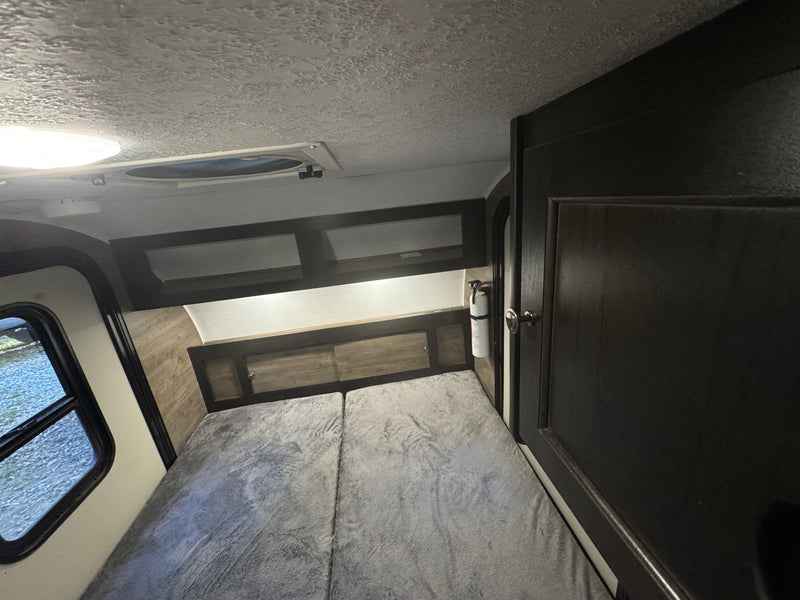 RV rental interior: cozy bedroom with a gray bed, dark overhead cabinets, a window, and a fire extinguisher. — photo 6