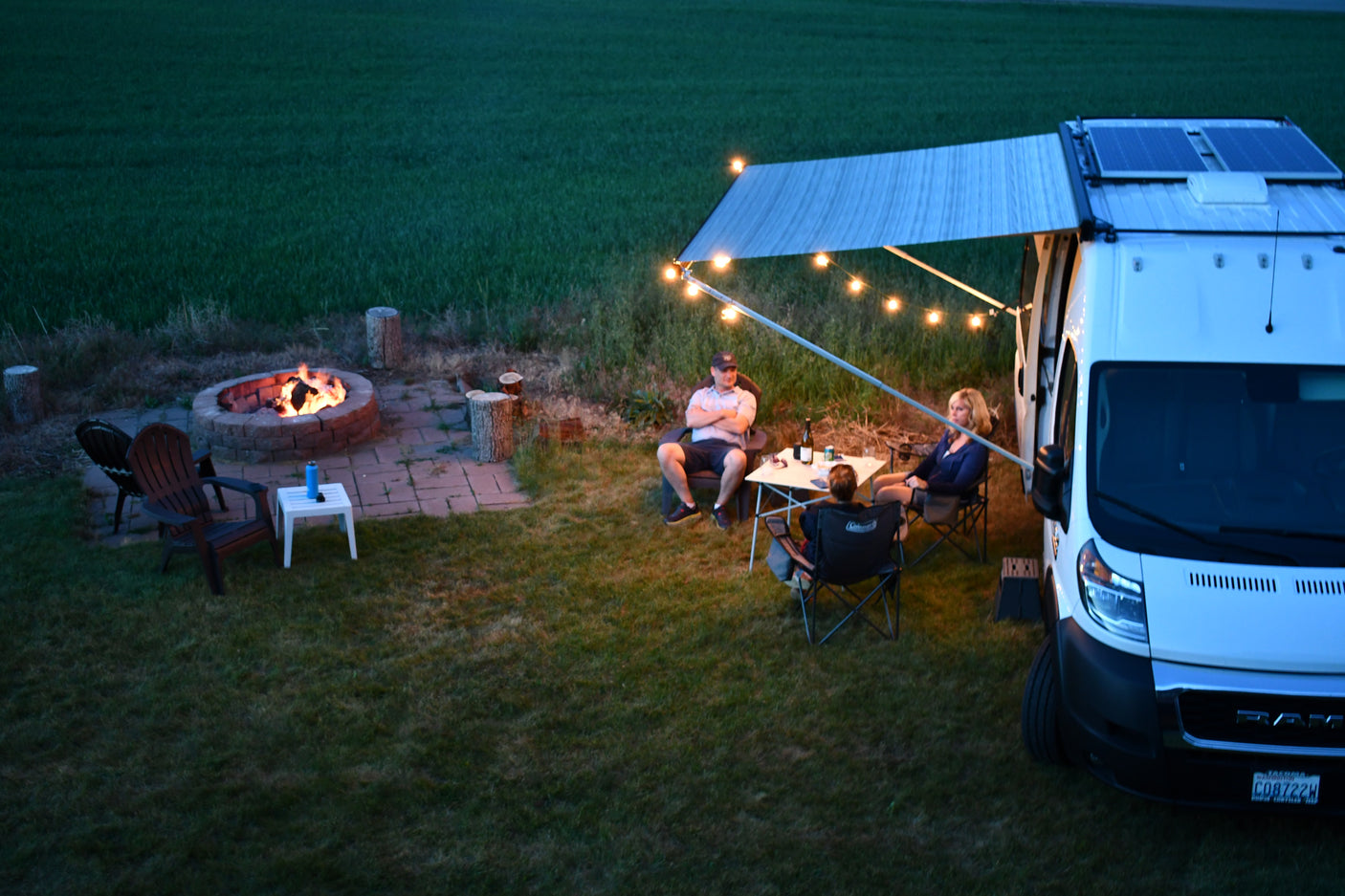 RV rental evening: A white camper van with an illuminated awning provides light for people relaxing around a table and campfire.