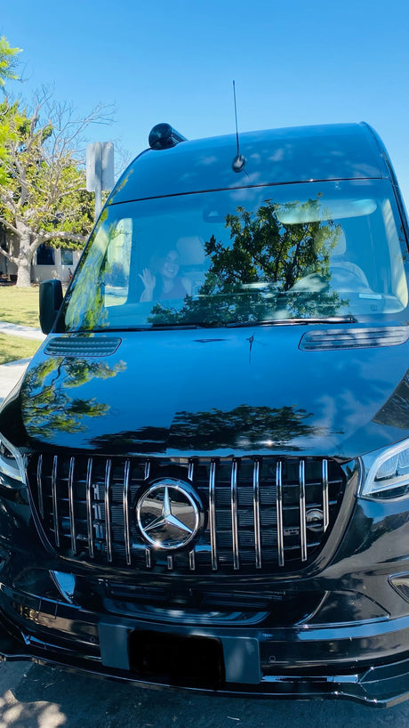 A glossy black Mercedes-Benz Sprinter van, likely an rv rental, with a person waving. Features a bold chrome grille and shiny reflections.