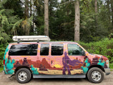 Striking rv rental van with a desert-themed mural (cowboy, cacti, sunset) and a rooftop cargo box, parked in a green forest.