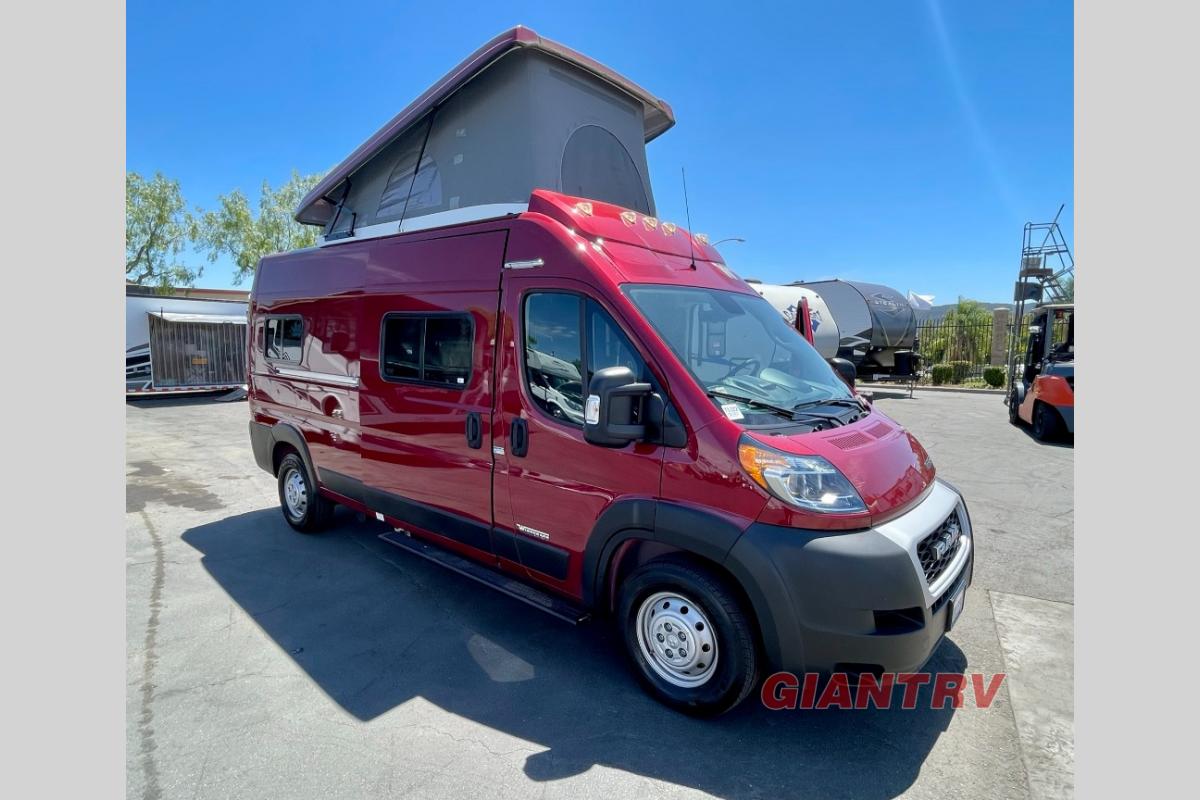  parked on asphalt with green foliage and a stone wall.|Stylish red Ram ProMaster RV rental parked curbside on a sunny day. Ready for adventure!|Experience an rv rental adventure in this red Winnebago Solis pop-top camper van — photo 3
