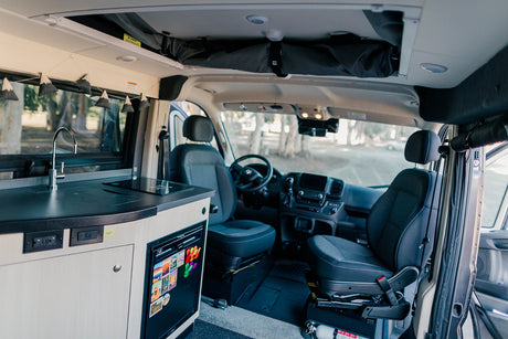 Inside an RV rental: a modern kitchenette with a sink and stovetop, a mini-fridge, front cabin seats, and overhead storage.