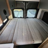 Comfortable grey upholstered bed in an RV rental camper van. The inviting sleeping area features large rear windows for light.