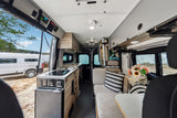 This RV rental showcases a modern camper van interior: kitchen with sink, comfy seating, overhead storage, and scenic outdoor views.