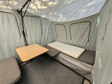 Inside an RV rental pop-up, showing a small table, grey cushioned seating, a foam bed, and carpeted floor.
