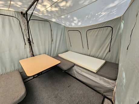Inside an RV rental pop-up, showing a small table, grey cushioned seating, a foam bed, and carpeted floor.