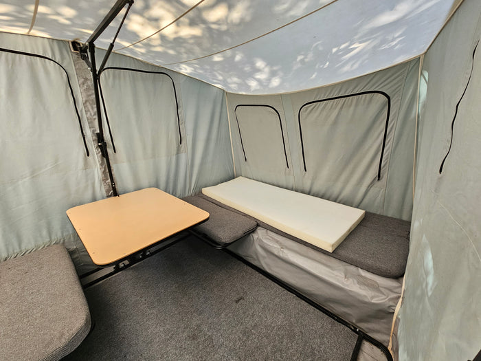 Inside an RV rental pop-up, showing a small table, grey cushioned seating, a foam bed, and carpeted floor.