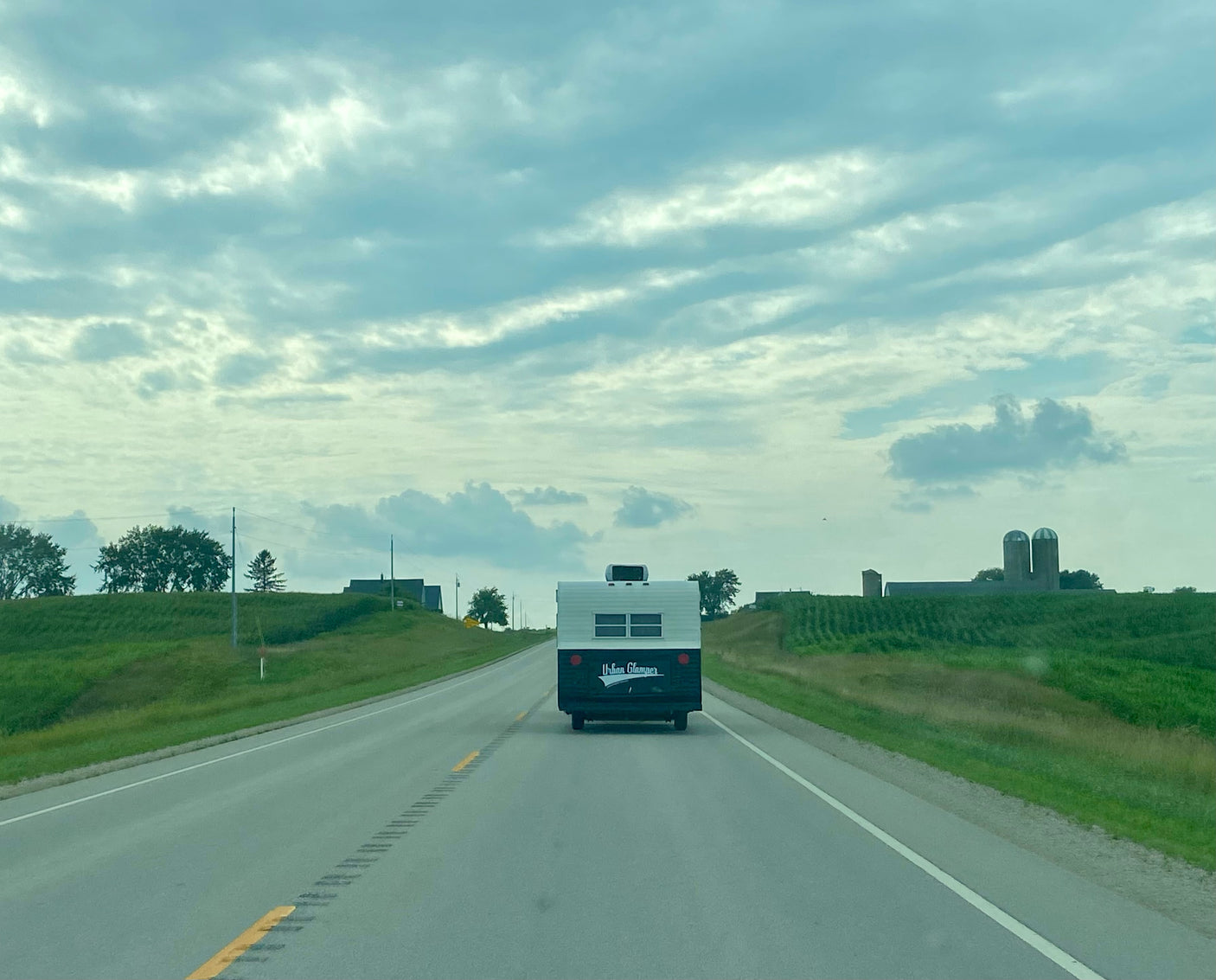 An 'Urban Glamper' RV rental on a rural highway. Green fields, distant silos, and cloudy skies define this road trip scene.