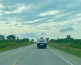 An 'Urban Glamper' RV rental on a rural highway. Green fields, distant silos, and cloudy skies define this road trip scene.